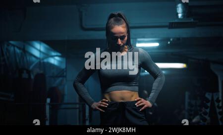 Donna forte con corpo atletico che indossa ascensori sportivi e fa esercizi vigorosi con il barbell vuoto in palestra buia con illuminazione a LED. L'atleta si allena prima della competizione sportiva. Allenamento CrossFit. Foto Stock