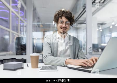 Giovane uomo d'affari dai capelli ricci in un abbigliamento elegante e casual che lavora diligentemente alla sua scrivania con laptop e cuffie. Foto Stock