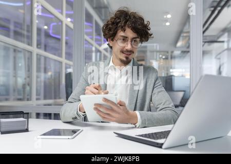 Giovane uomo professionista con capelli ricci che lavora al notebook in un ufficio aziendale, mostrando attenzione e competenza. Foto Stock