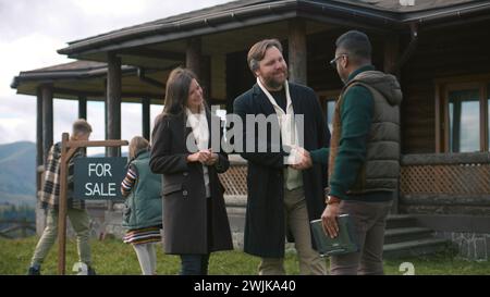 L'agente immobiliare afroamericano dà le chiavi alla felice coppia caucasica e si congratula con l'acquisto. Le famiglie con bambini acquistano una casa moderna ed elegante in montagna. Proprietà residenziale in vendita. Foto Stock