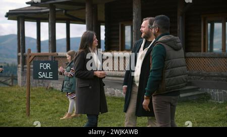 L'agente immobiliare afroamericano dà le chiavi alla felice coppia caucasica e si congratula con l'acquisto. Le famiglie con bambini acquistano una casa moderna ed elegante in montagna. Proprietà residenziale in vendita. Foto Stock