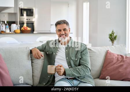 Uomo di mezza età felice che tiene una tazza di caffè rilassante sul divano a casa. Verticale. Foto Stock