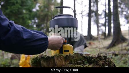 Il bollitore turistico si trova su una stufa a gas portatile da campeggio. Il viaggiatore maschile accende il bruciatore a gas. Un gruppo di amici escursionisti si è fermato per riposarsi dopo aver fatto trekking in montagna. Scoperta della natura e concetto di svago attivo. Foto Stock