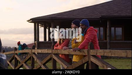 Un gruppo di turisti multietnici si trova vicino a una recinzione di legno, parla e guarda il bellissimo tramonto. Gli amici escursionisti si rilassano durante le vacanze in montagna. Elegante cottage in legno sullo sfondo. Al rallentatore. Foto Stock