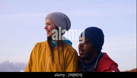 Felice coppia multietnica sorride e abbraccia, guarda lo splendido tramonto godendosi la natura durante il viaggio in campagna. La giovane famiglia di viaggiatori riposa dopo un'escursione in montagna. Al rallentatore. Foto Stock