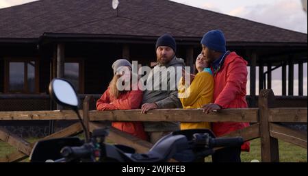 Un gruppo di turisti multietnici si trova vicino a una recinzione di legno, parla e guarda il bellissimo tramonto. Gli amici escursionisti si rilassano durante le vacanze in montagna. Elegante cottage in legno sullo sfondo. Al rallentatore. Foto Stock