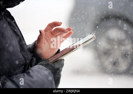 Mani femminili con smartphone primo piano su sfondo sfocato dell'auto durante le nevicate. Donna che usa il cellulare in una strada di città in inverno Foto Stock