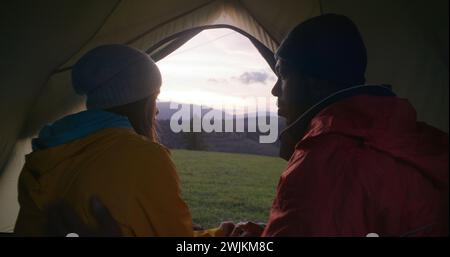 Una coppia multietnica di escursionisti siede in tenda sulla cima della collina, bacia e guarda al bellissimo tramonto. Un uomo afroamericano e una donna caucasica si godono gli incredibili paesaggi delle montagne durante il viaggio. Appassionati di attività all'aria aperta. Foto Stock