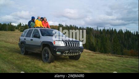 Gruppi di turisti multietnici insieme viaggiano in vacanza in montagna. Gli escursionisti che si alzano dal tetto dell'auto, ridono e sbattono le mani durante il viaggio. La gente ama guidare nel portello. Appassionati di attività all'aria aperta Foto Stock