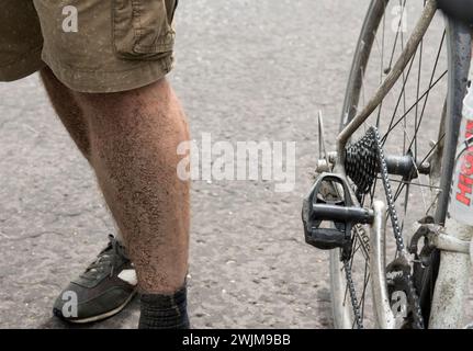 Le zampe fangose di un uomo che indossa pantaloncini corti e calze accanto a una bicicletta durante un giro in bicicletta. Foto Stock