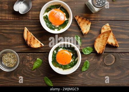 Uova en cocotte (uova al forno) con panna, сheese e spinaci in porzione recipiente di ceramica su fondo di legno. Stile rustico, vista dall'alto, disposizione piatta Foto Stock