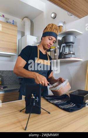 immagine verticale del giovane latino in uniforme da chef a casa felice di preparare la torta per la sua famiglia guardando video tutorial con il telefono. Foto Stock