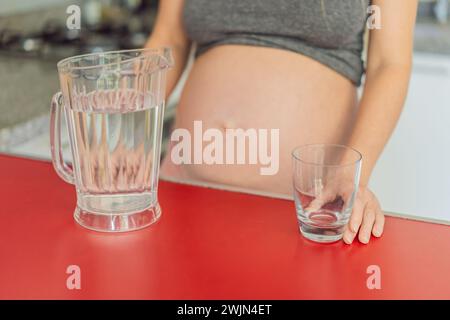 Abbracciando i benefici vitali dell'acqua durante la gravidanza, una donna incinta sta in cucina con un bicchiere, evidenziando il ruolo cruciale dell'idratazione in Foto Stock