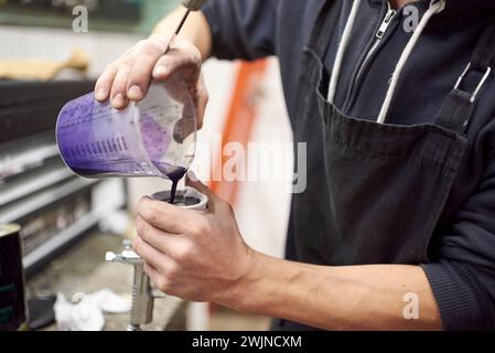 Persona irriconoscibile che versa vernice viola in un serbatoio della pistola a spruzzo nella sua officina. Persone vere che lavorano. Foto Stock