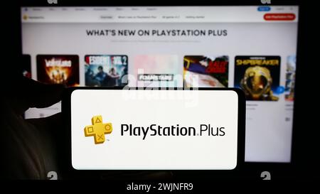 Persona che detiene un cellulare con il logo del servizio di abbonamento PlayStation Plus (PS Plus) di fronte alla pagina Web aziendale. Mettere a fuoco il display del telefono. Foto Stock