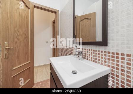 Un piccolo bagno con piastrelle simili a gresite, mobili in legno scuro, uno specchio con una cornice coordinata e un lavandino in porcellana bianca Foto Stock