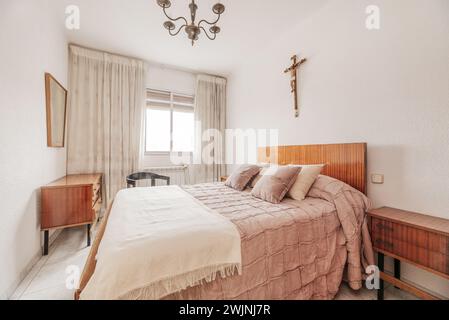 Camera da letto con testiera e mobili in legno verniciato, copriletto e cuscini coordinati, una croce cattolica sulle pareti e una finestra con tende Foto Stock