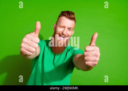 Foto di un uomo soddisfatto con barba lunga allo zenzero elegante t-shirt con un occhio lampeggiante che mostra i pollici isolati su uno sfondo di colore verde Foto Stock