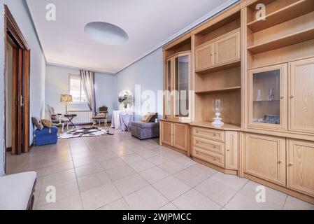 Immagine frontale di un soggiorno con una libreria di legno chiaro, una poltrona quadrata grigia e diverse poltrone sotto una finestra Foto Stock