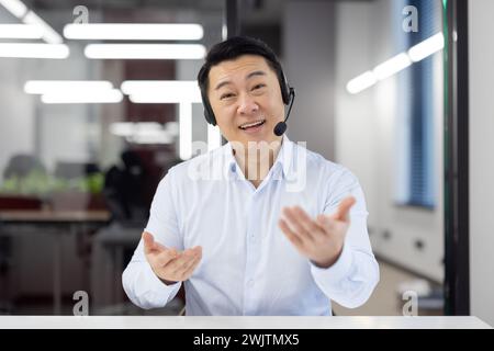 Un allegro uomo d'affari asiatico con cuffie e auricolari impegnato attivamente in una conversazione in ufficio. Foto Stock