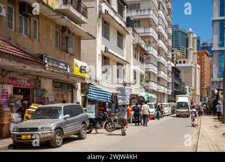 Un portiere porta in testa dei rotoli di carta igienica, passando davanti ai negozi in questa visione generale della vita in una strada nel centro di Dar es Salaam, Tanzania. Foto Stock