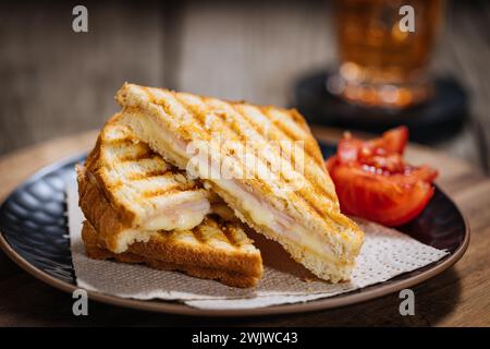 Toast tagliati a fette con formaggio Gouda fuso e prosciutto di tacchino su fondo di legno con pomodoro ciliegino su un piatto nero. Servito con tè freddo. Foto Stock