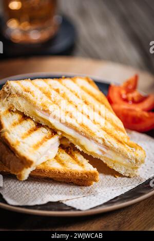 Toast tagliati a fette con formaggio Gouda fuso e prosciutto di tacchino su fondo di legno con pomodoro ciliegino su un piatto nero. Servito con tè freddo. Foto Stock