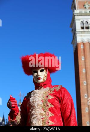 Persona mascherata con volto coperto da maschera e grande copricapo rosso con guanti a mano durante il carnevale di Venezia con il campanile di San Marco nel backgrou Foto Stock