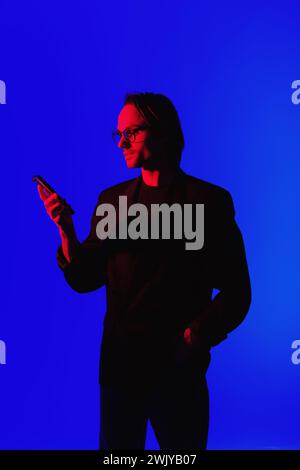 Ritratto di un uomo elegante con occhiali da vista che utilizza uno smartphone con luce rossa al neon su uno sfondo blu brillante Foto Stock