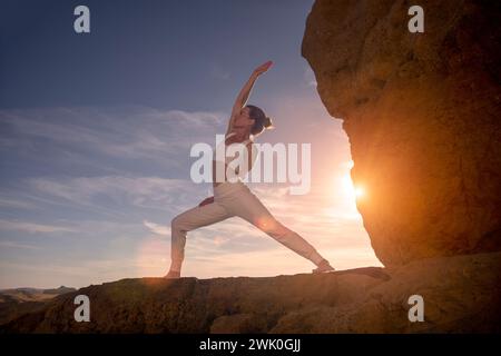 donna in piedi sulle rocce all'alba, al tramonto mentre fa una posa da yoga da guerriero al contrario. Foto Stock