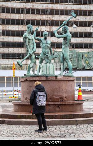 Donna con uno zaino davanti alla statua dei tre fabbri e edificio per uffici in demolizione sullo sfondo a Helsinki, Finlandia Foto Stock