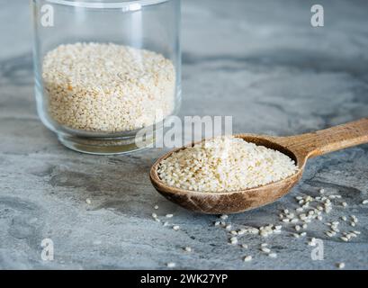 Vaso di vetro e cucchiaio di legno riempito con semi di sesamo su un piano di lavoro in pietra Foto Stock