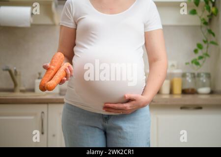 Una donna incinta tiene le carote tra le mani mentre sta in piedi nella sua cucina di casa. Gravidanza e corretta alimentazione mentre si aspetta un bambino Foto Stock