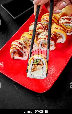 Primo piano della mano femminile con le bacchette per raccogliere appetitosi sushi roll ripieni di anguilla, cetriolo e formaggio spalmabile conditi con cheddar da VIBRANT Foto Stock