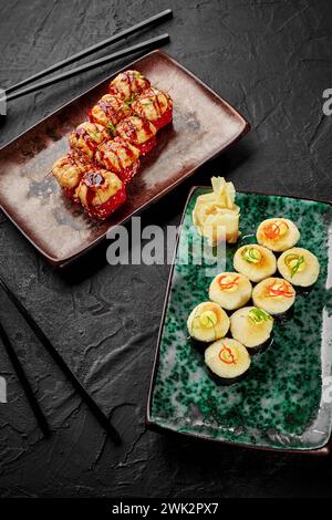 Due tipi di appetitosi sushi panini al forno con cappelli di formaggio dorato morbido guarniti con maionese piccante, scaglie di verdure e fili di peperoncino serviti sul colore Foto Stock