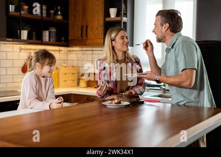 Una scena emozionante si svolge mentre una famiglia assaggia insieme una deliziosa torta al cioccolato nel calore della sua cucina soleggiata, condividendo sorrisi e sorrisi Foto Stock