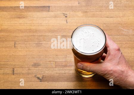 Mano di un adulto che afferra saldamente una pinta di vetro trasparente riempita di birra color ambra su un tavolo in legno caldo. La testa bianca spumeggiante suggerisce la birra i Foto Stock