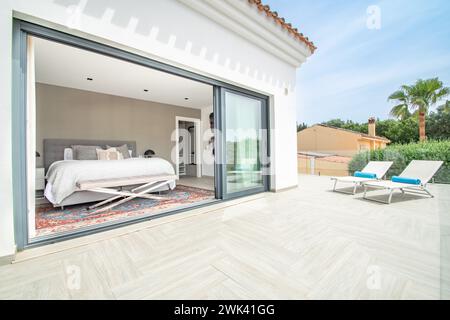 una lussuosa camera da letto padronale con porte di balcone aperte che conducono alla terrazza solarium con lettini nella villa mediterranea Foto Stock