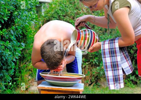 Un ragazzo si lava la faccia in giardino sopra una ciotola e sua sorella gli versa l'acqua da una brocca, tenendo un asciugamano in mano. Foto Stock