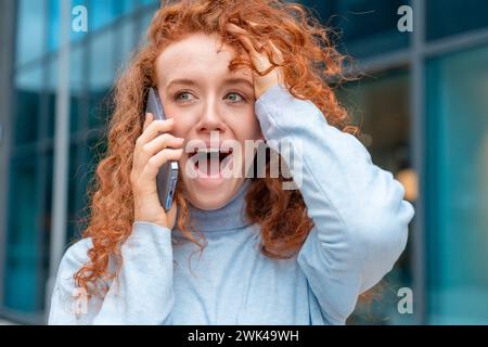 una donna con una camicia blu che parla al telefono cellulare e ha scioccato le notizie. Tenendo la mano sulla testa e il telefono, chiudile l'orecchio. Stare da soli fuori. Busi Foto Stock