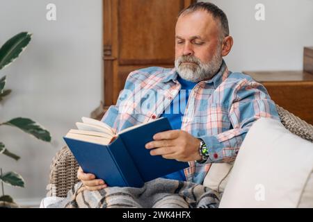 Sorridente anziano anziano uomo maturo che ride leggendo un libro riposato, seduto sul divano nel soggiorno Foto Stock