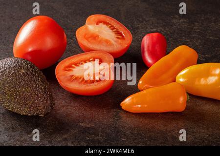 Un mix vibrante di pomodori rossi freschi, peperoni verdi e avocado maturo disposti su un elegante tavolo nero. Questa foto dai toni bassi mostra la colorata A. Foto Stock