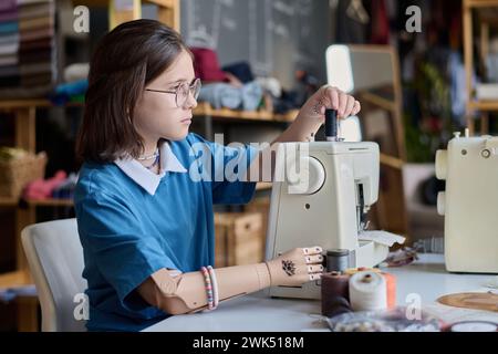 Vista laterale ritratto di adolescente con la mano protesica che utilizza una macchina da cucire nello spazio di copia del corso di formazione professionale Foto Stock