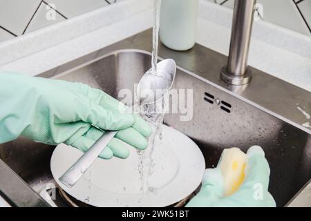 Mani in guanti verdi lavando un cucchiaio sull'acqua corrente in un lavello da cucina in acciaio inox, pila di piastre sporche. dispenser di detergente Foto Stock