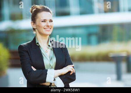 lavoratrice moderna e felice di mezza età vicino all'edificio degli uffici in giacca nera con braccia incrociate. Foto Stock