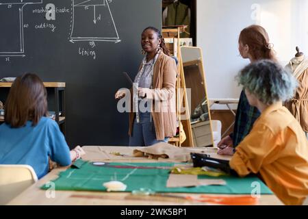 Ritratto di una maestra afroamericana in piedi a bordo che spiega la produzione di abbigliamento a un gruppo di ragazze nello spazio copia laboratorio dell'atelier Foto Stock
