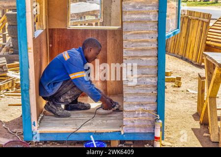 falegname afroamericano che crea una casa wendy, capannone di legno all'aperto, da vicino, sta macinando le tavole e assemblandole in pannelli per il pavimento Foto Stock