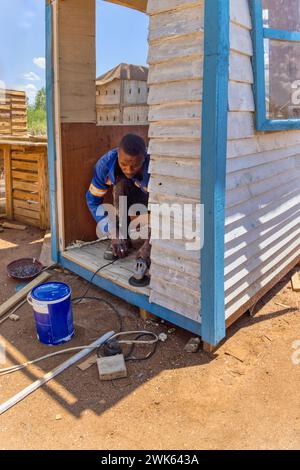 lavoratore afroamericano che realizza una casa wendy, capannone di legno all'aperto, sta usando una smerigliatrice angolare per levigare il pavimento Foto Stock