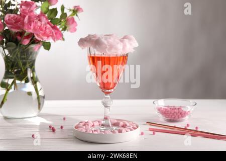 Cocktail di caramelle di cotone in vetro, marshmallow, vaso con rose rosa e cannucce su un tavolo di legno bianco su sfondo grigio Foto Stock