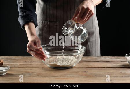 Fare il pane. Donna che versa l'acqua nella ciotola con la farina al tavolo di legno, primo piano Foto Stock